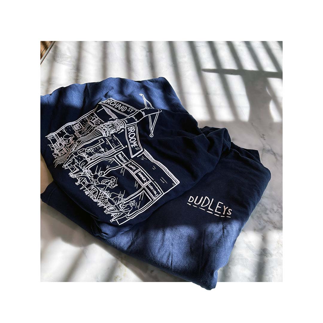 Navy Bella Canvas tri-blend shirts with custom Dudleys restaurant artwork, featuring a detailed line drawing and logo, folded on a marble surface with shadows