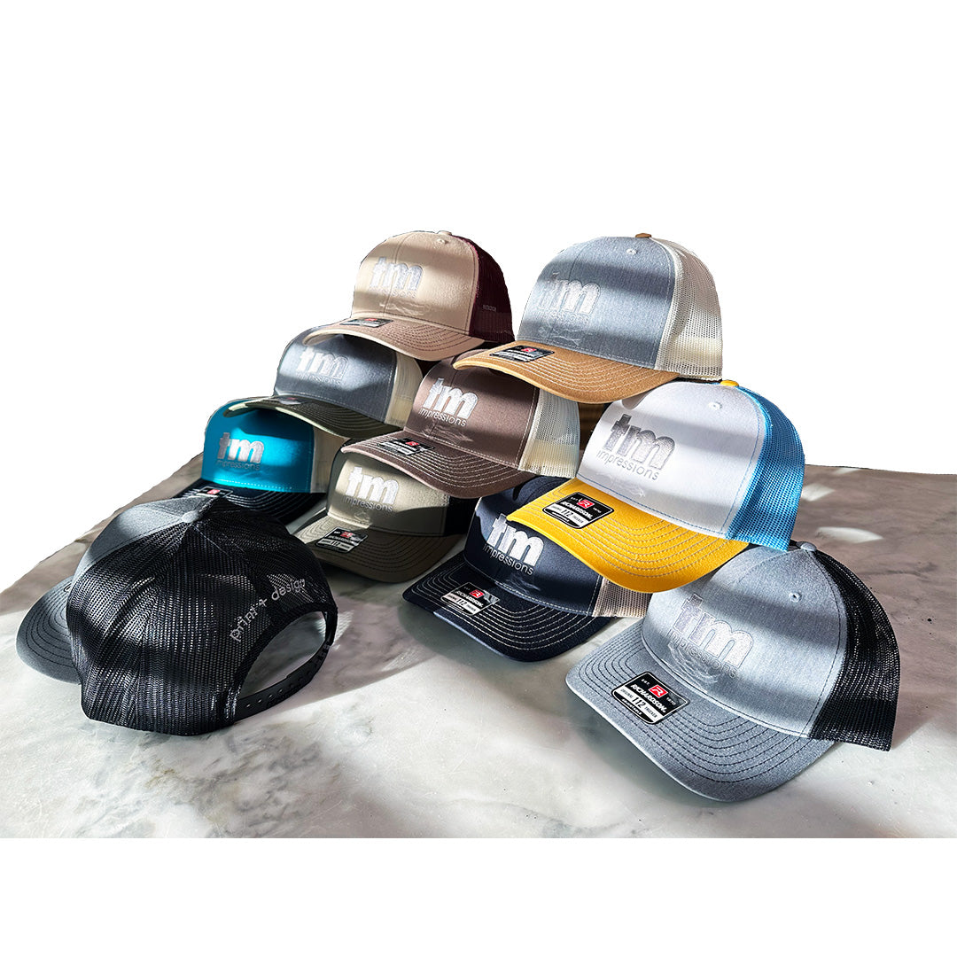 Multiple Richardson 112 hats with TM Impressions logo embroidery in various colors including teal, gold, black, and gray, arranged in a pyramid display on a marble surface