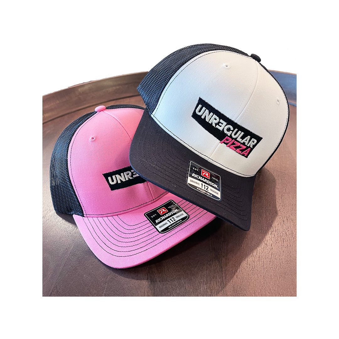 Two Richardson 112 trucker hats with Unregular Pizza embroidery—one in pink, one in white with black mesh back—displayed on a wooden table