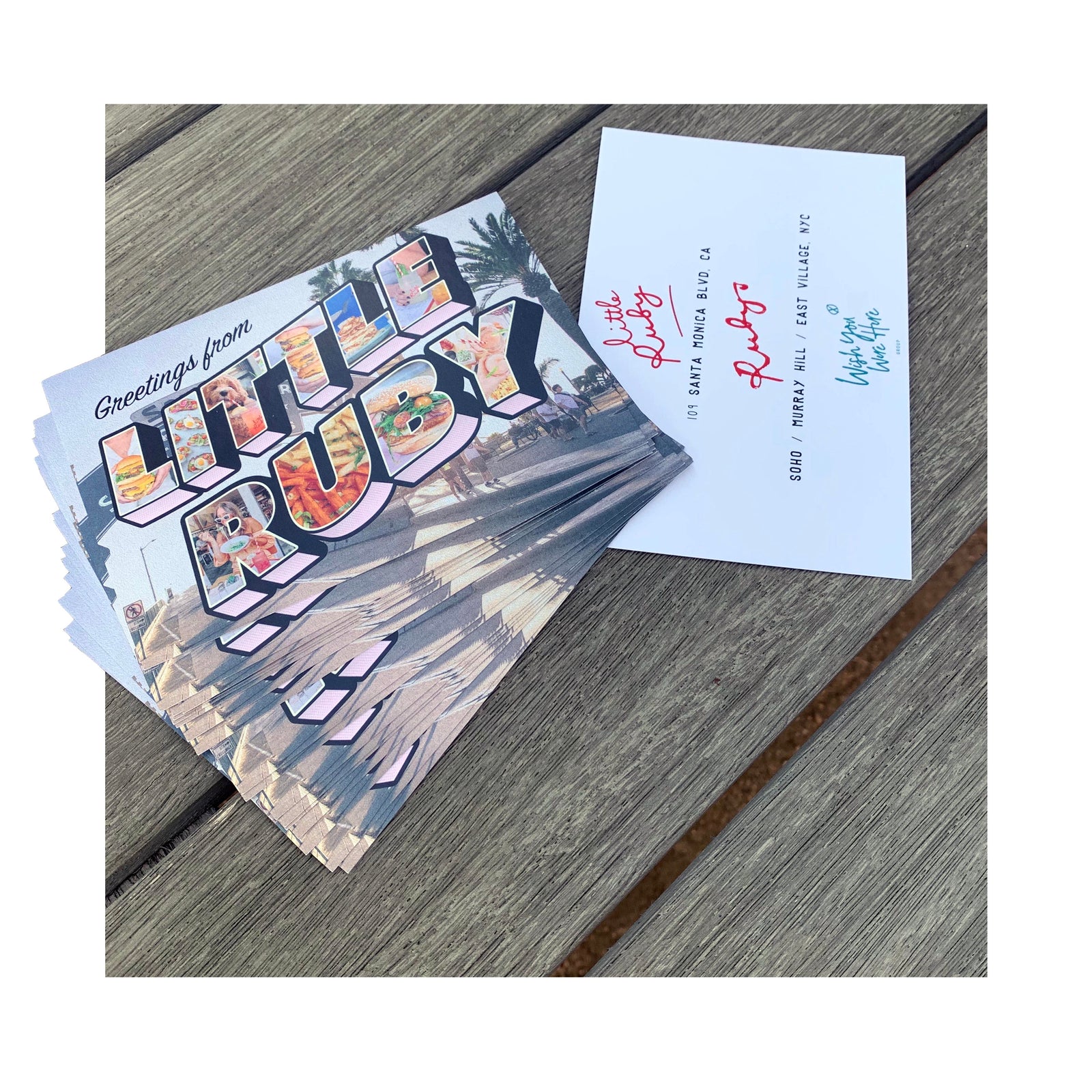 Stack of custom “Greetings from Little Ruby” postcards with vibrant beach imagery and branded back design, displayed on a wooden surface