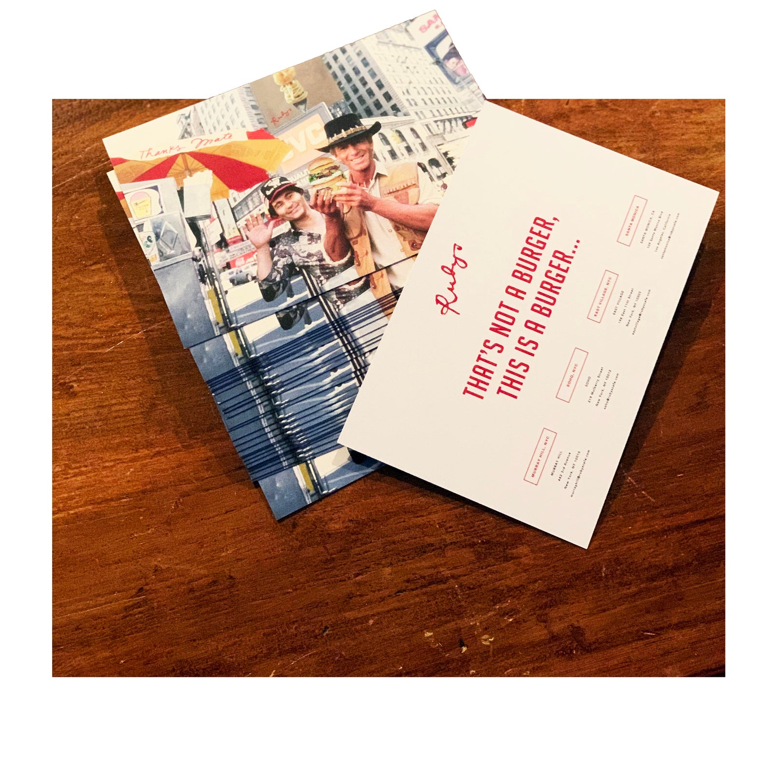 Stack of postcards on a wooden table featuring a colorful street scene with two men posing in front of a NYC hot dog cart. The reverse side of the top postcard reads “THAT’S NOT A BURGER, THIS IS A BURGER…” in bold red text, with restaurant info and Ruby’s branding.