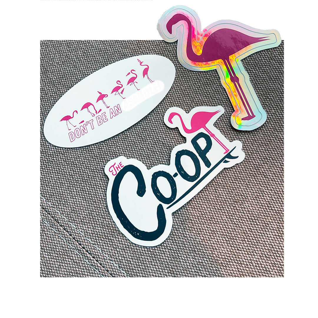 Assorted custom printed stickers for The Co-Op and a flamingo-themed brand, shown on textured fabric including holographic and oval styles