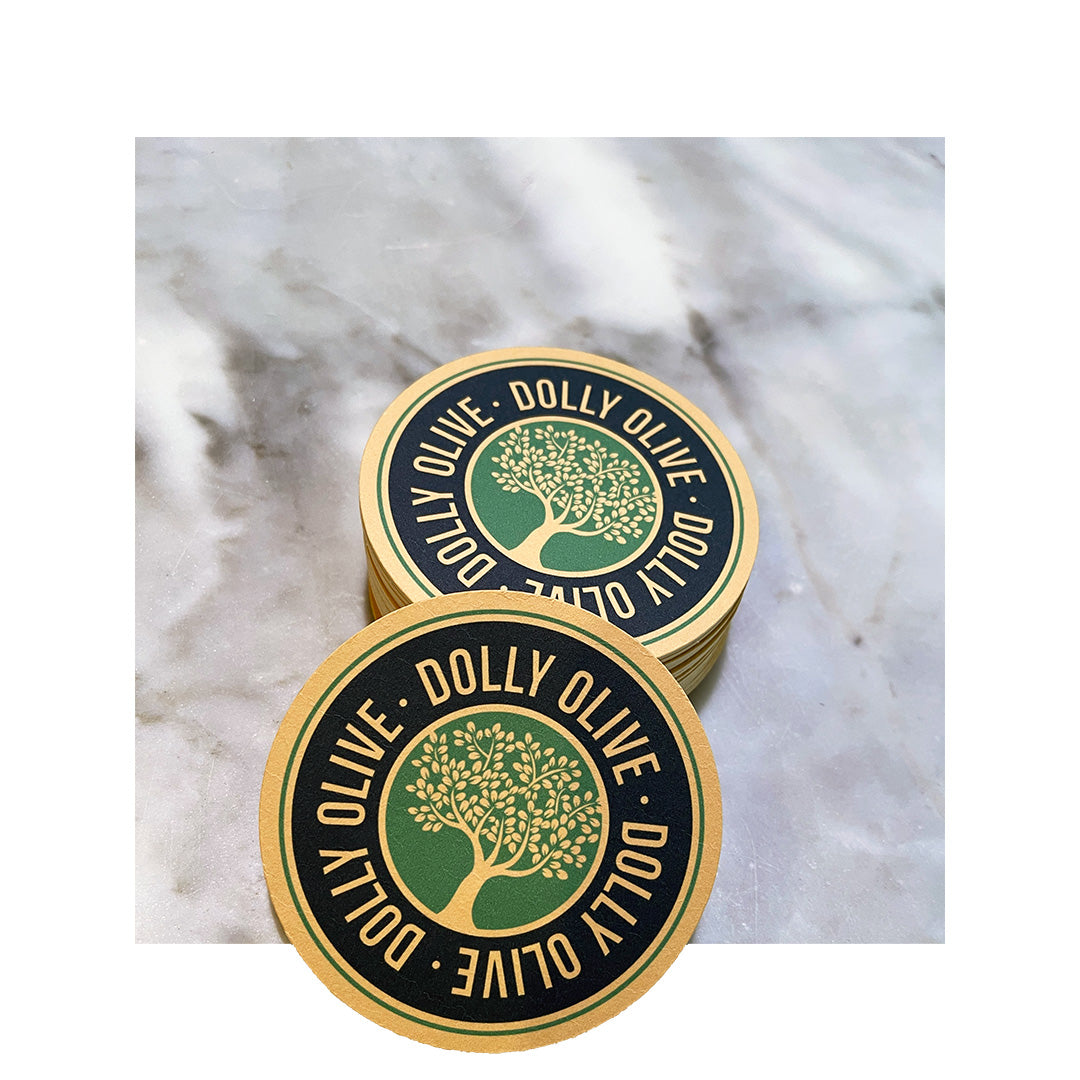 Round coasters with a bold “Dolly Olive” logo surrounding a stylized green tree on a black and tan background, stacked on a marble surface