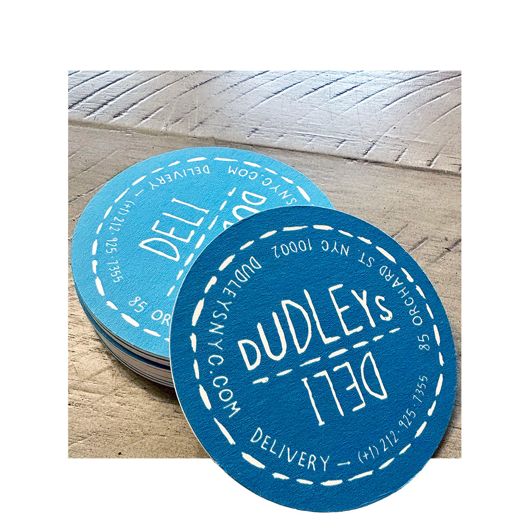 Stack of round blue coasters with white stitched design and “Dudley’s Deli” branding, placed on a rustic wood surface