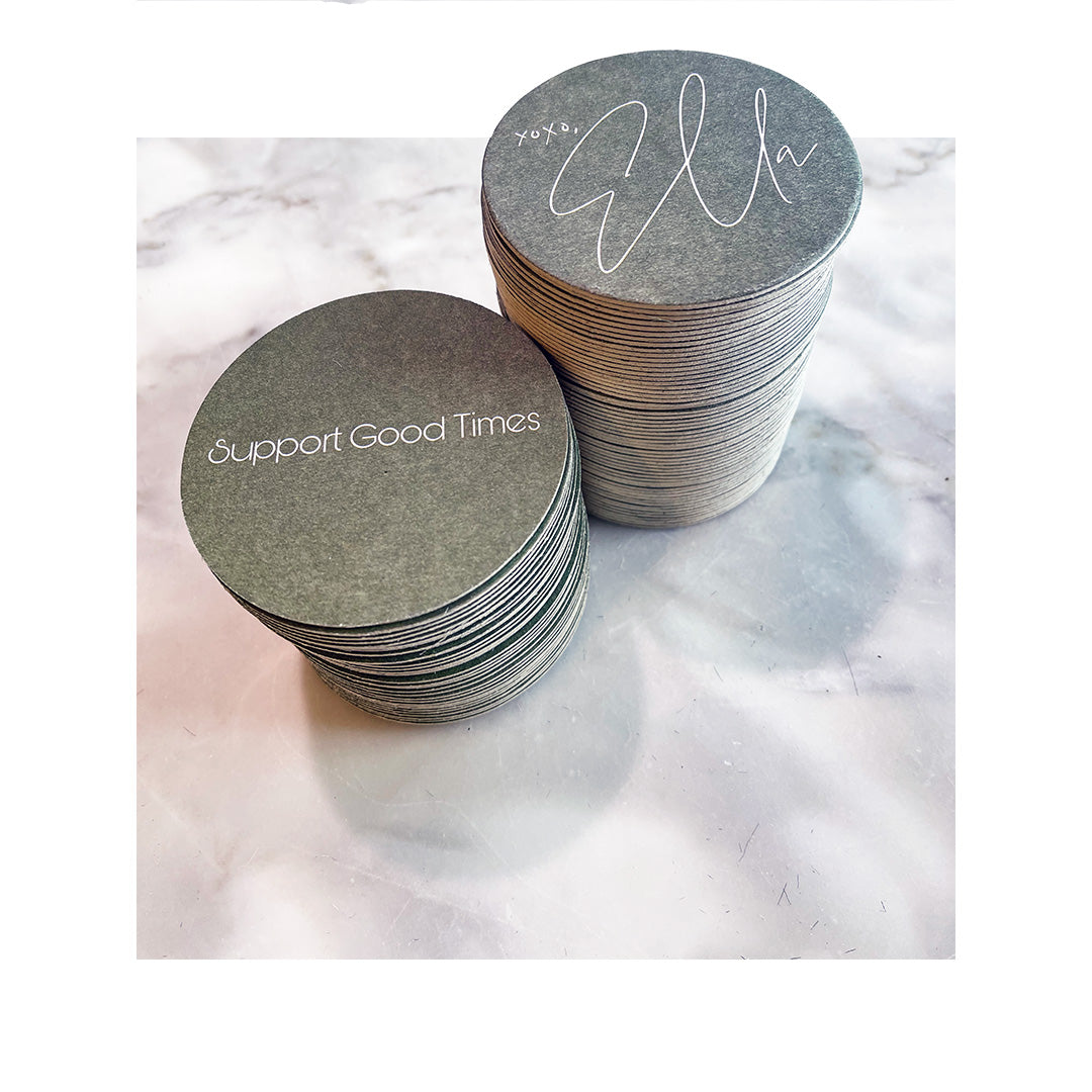 Two stacks of round coasters on a marble surface, one reading “Support Good Times” and the other featuring a white cursive signature-style design that says “XOXO, Ella.”