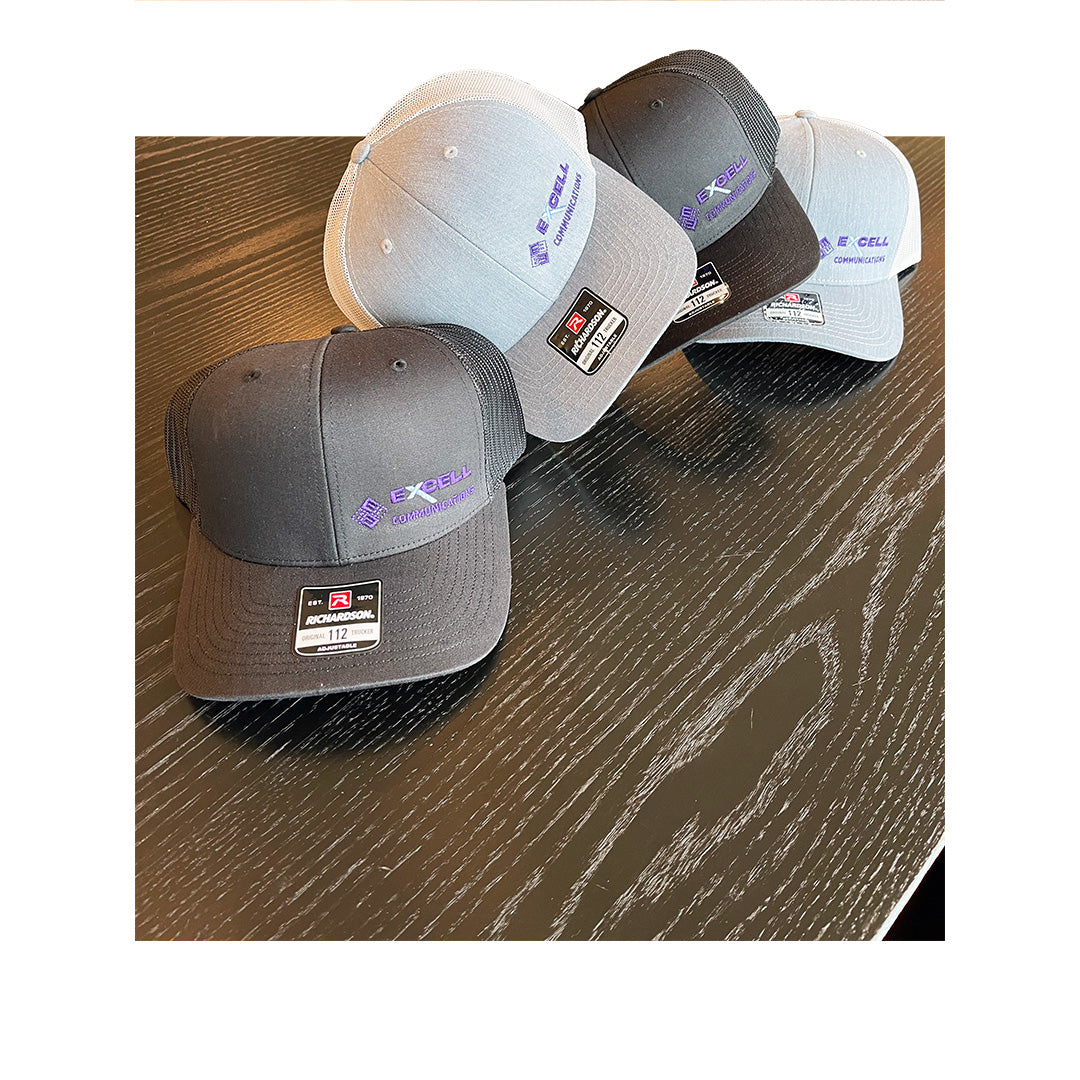 Three gray and black Richardson 112 hats with Excell Communications logo embroidered in purple, arranged diagonally on a black wood table