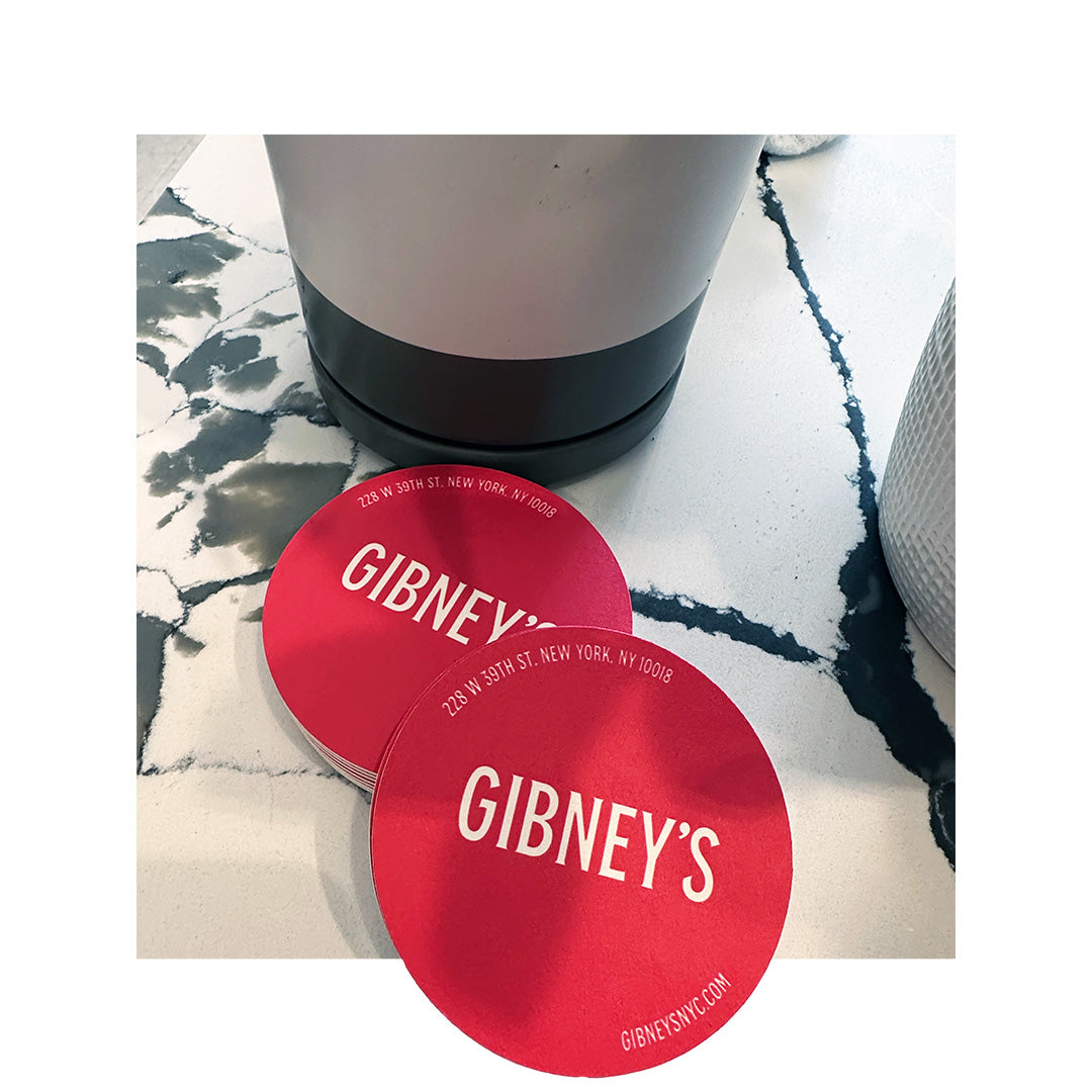 Stack of bright red round coasters printed with “Gibney’s” and the NYC address in white text, placed on a marble table next to a modern drink dispenser.