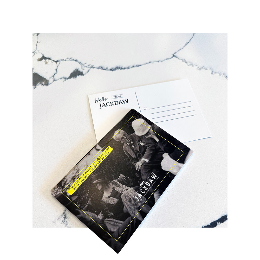 Postcard with a vintage photo and yellow text border featuring the JACKDAW logo, placed on a marble surface beside the back side of the postcard, which reads “Hello from JACKDAW” with space to write a message and address
