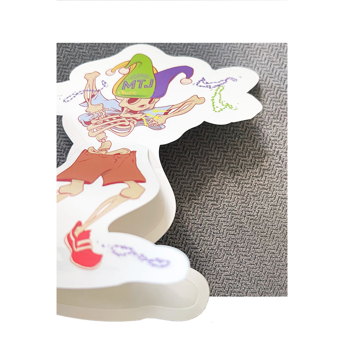 Close-up of a custom die-cut sticker showing a skeleton riding a unicycle with “MTJ” on a green and purple hat, placed on textured fabric