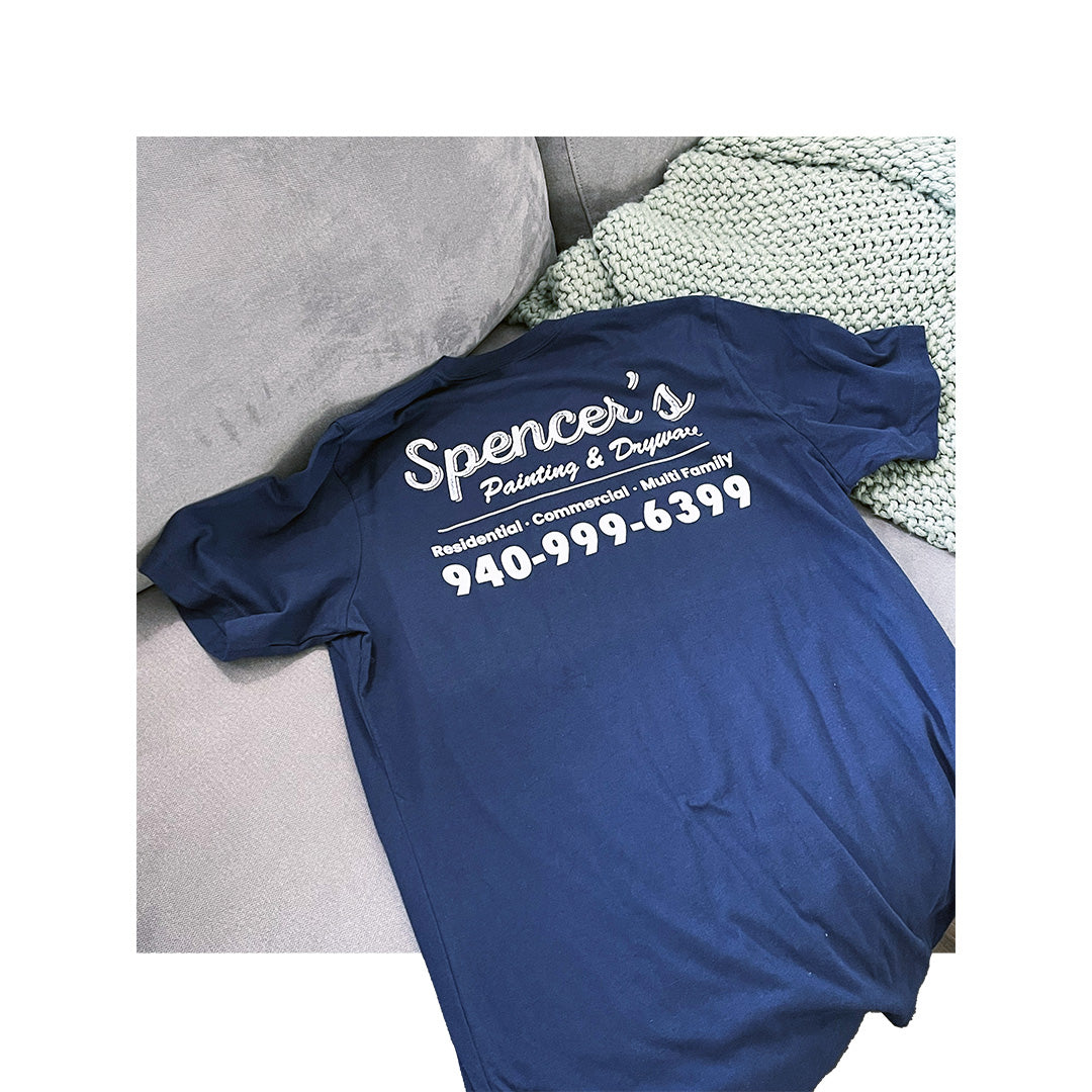 Navy Bella Canvas 3001 t-shirt with Spencer's Painting & Drywall business information and phone number printed in white on the back, displayed on a couch