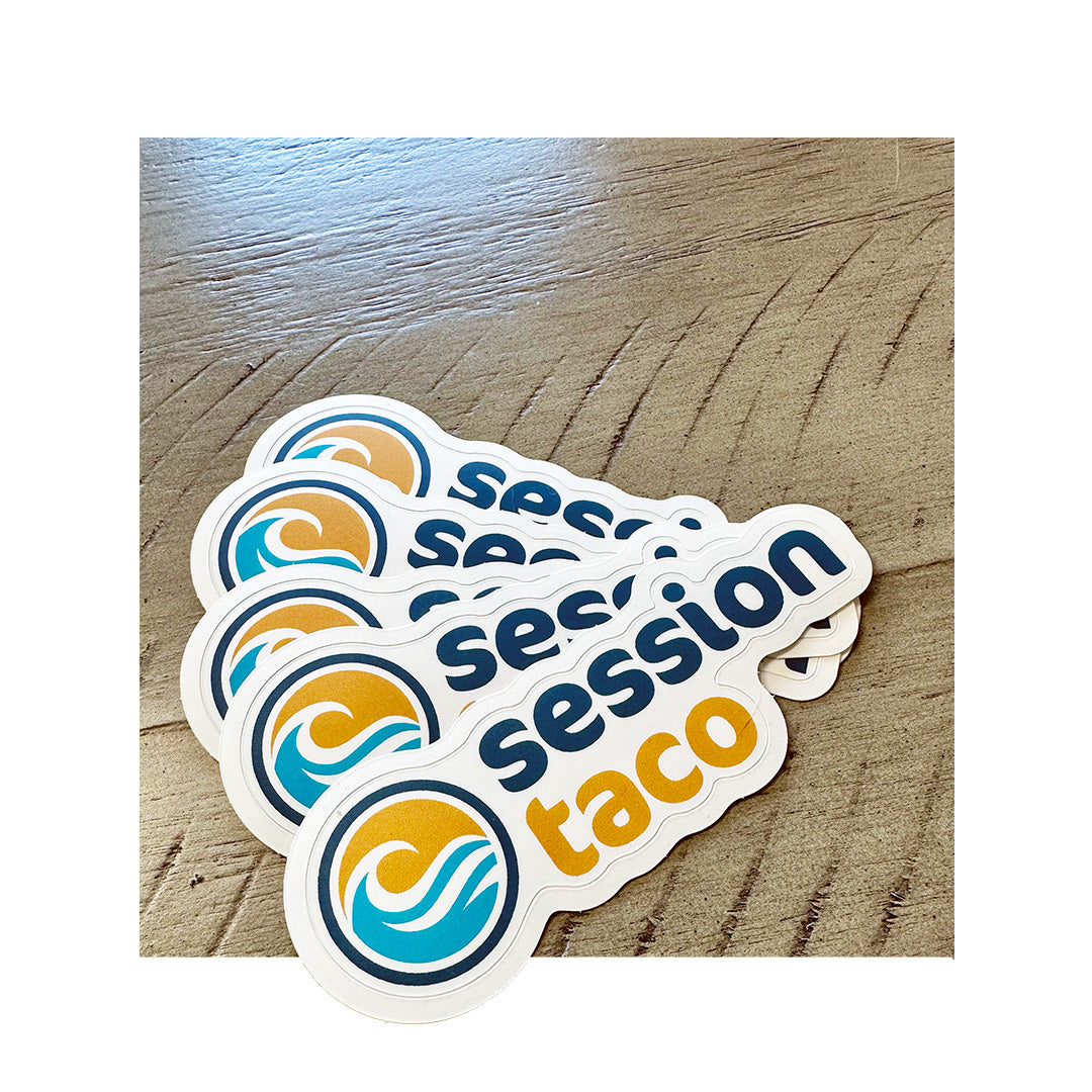 Custom die-cut stickers for Session Taco featuring a wave and sun logo in teal and orange colors, placed on a wooden surface