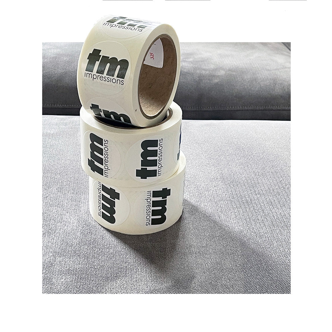 Stacked rolls of custom circle labels with TM Impressions logo, printed on white BOPP material with gloss finish