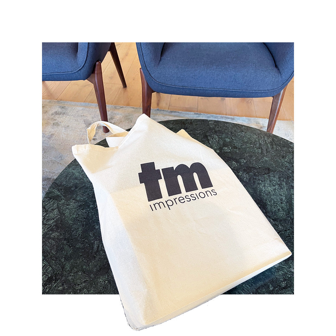 Canvas tote bag printed with 'tm impressions' logo in black ink, styled on a modern table