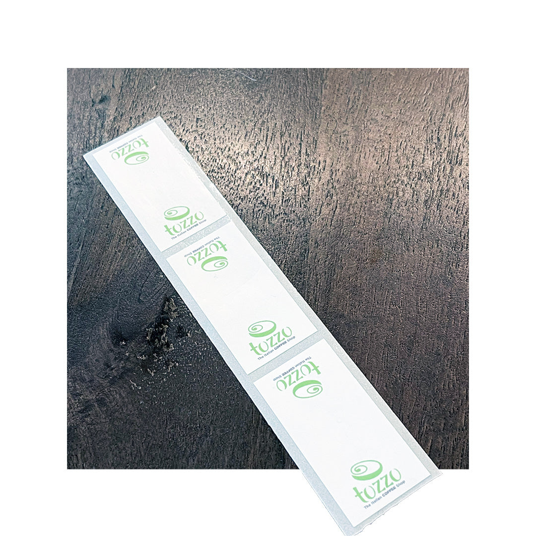 Strip of custom rectangle labels for Tozzo, printed on white BOPP with green logo and text, shown on dark wood surface