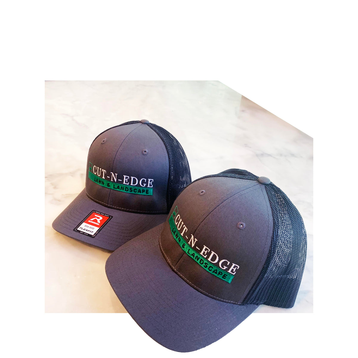 Two dark gray Richardson 112 snapback hats with custom Cut-N-Edge Lawn & Landscape embroidery on a marble background