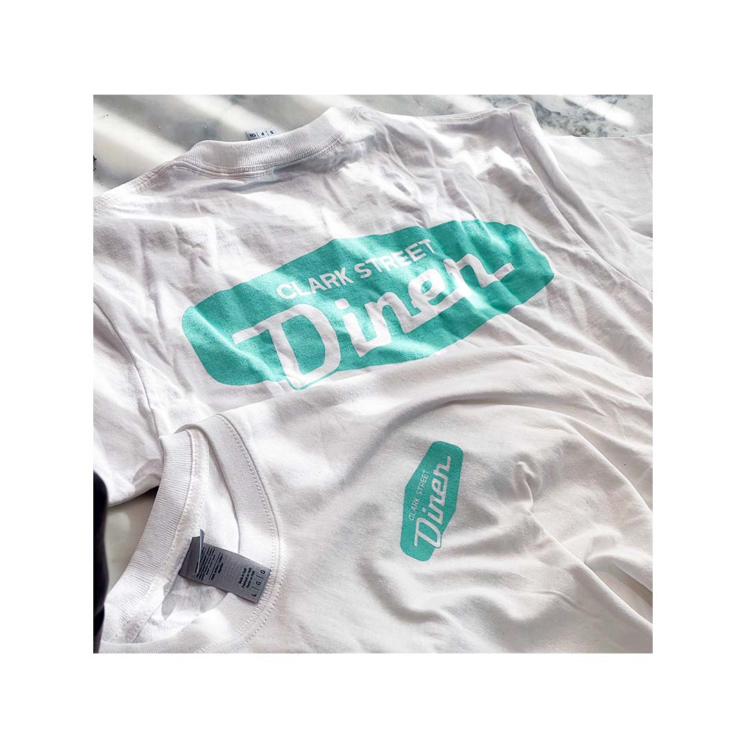 White Gildan 2000 T-shirts with teal front and back logo prints for Clark Street Diner, laid flat in natural light