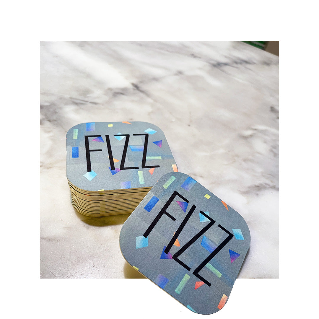 Stack of square coasters with rounded corners featuring a bold “FIZZ” design and colorful confetti-style graphics, placed on a marble surface