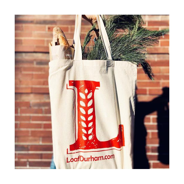 Canvas tote bag printed with large red 'L' and LoafDurham.com logo, filled with bread and greenery
