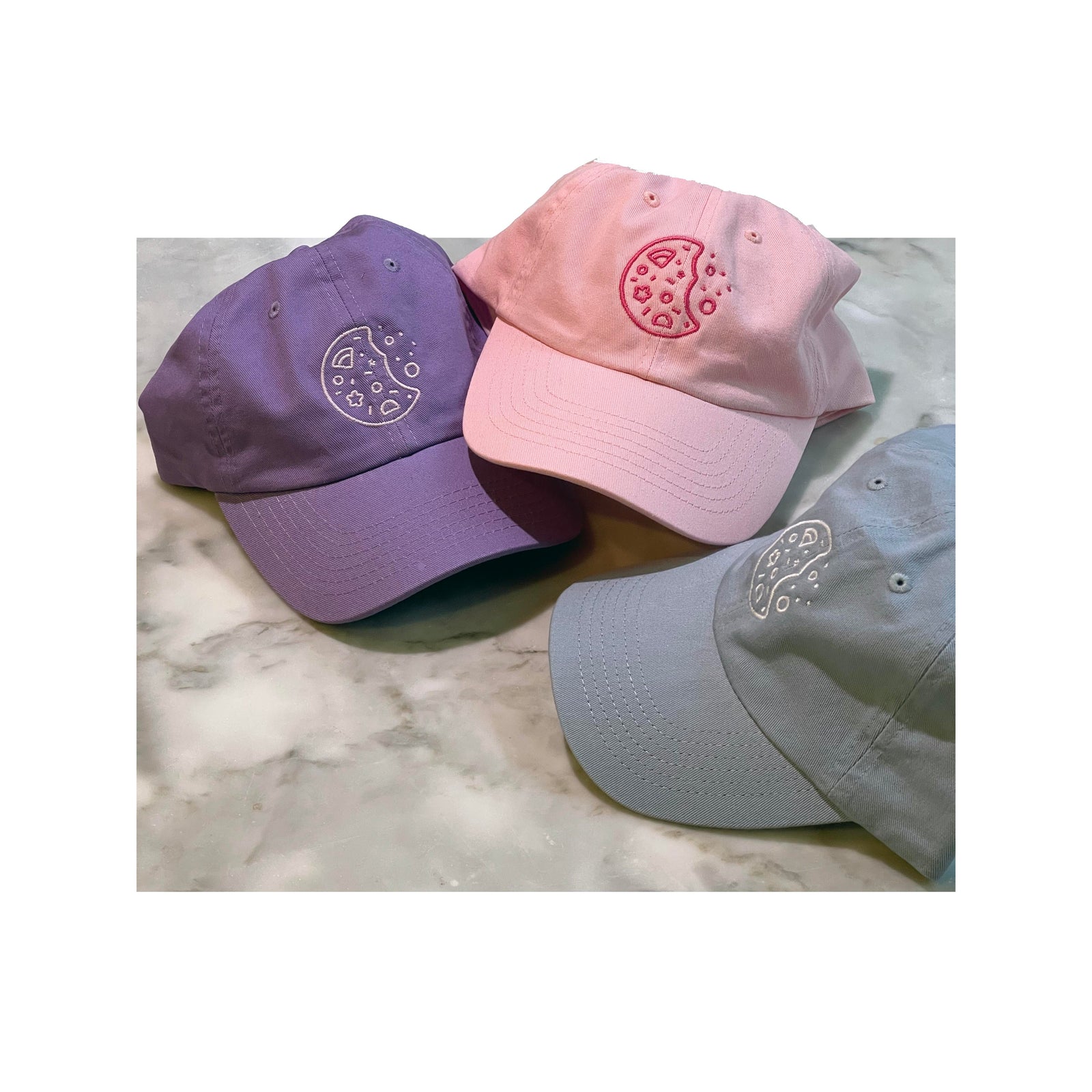 Pastel dad hats in lavender, pink, and light blue with minimalist cookie embroidery on each front pane