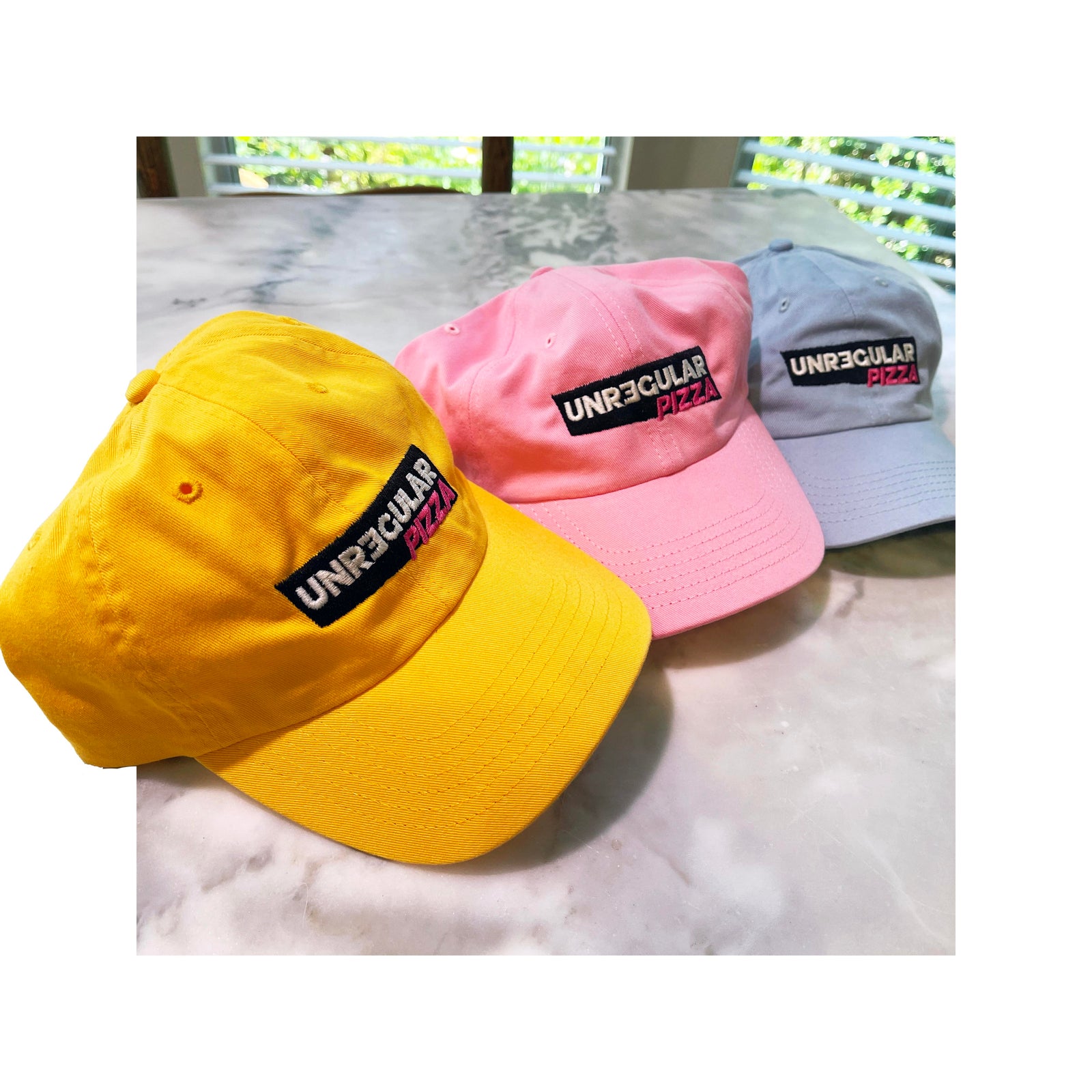 Bright yellow, pink, and blue "Unregular Pizza" embroidered dad hats on marble – Valucap VC300a