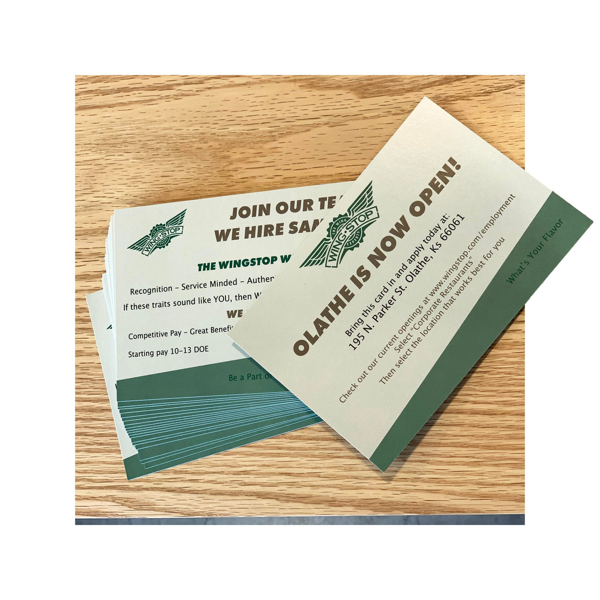 Stack of custom Wingstop postcards promoting a new store opening in Olathe, Kansas, with bold green branding and hiring callouts displayed on a light wood surface