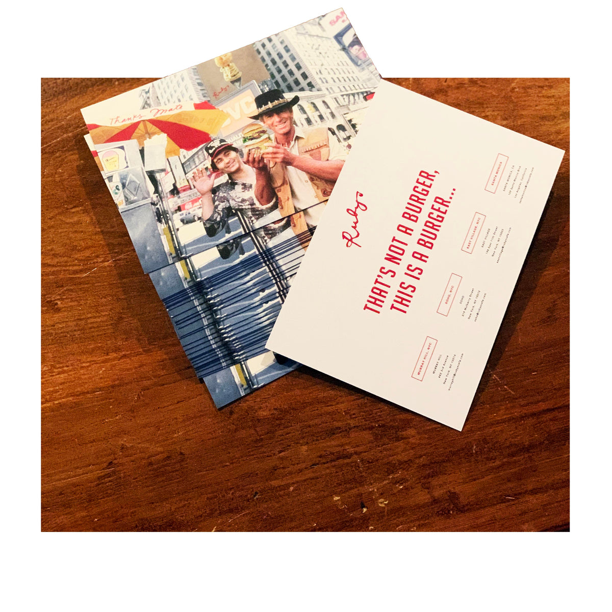 Stack of postcards on a wooden table featuring a colorful street scene with two men posing in front of a NYC hot dog cart. The reverse side of the top postcard reads “THAT’S NOT A BURGER, THIS IS A BURGER…” in bold red text, with restaurant info and Ruby’s branding.